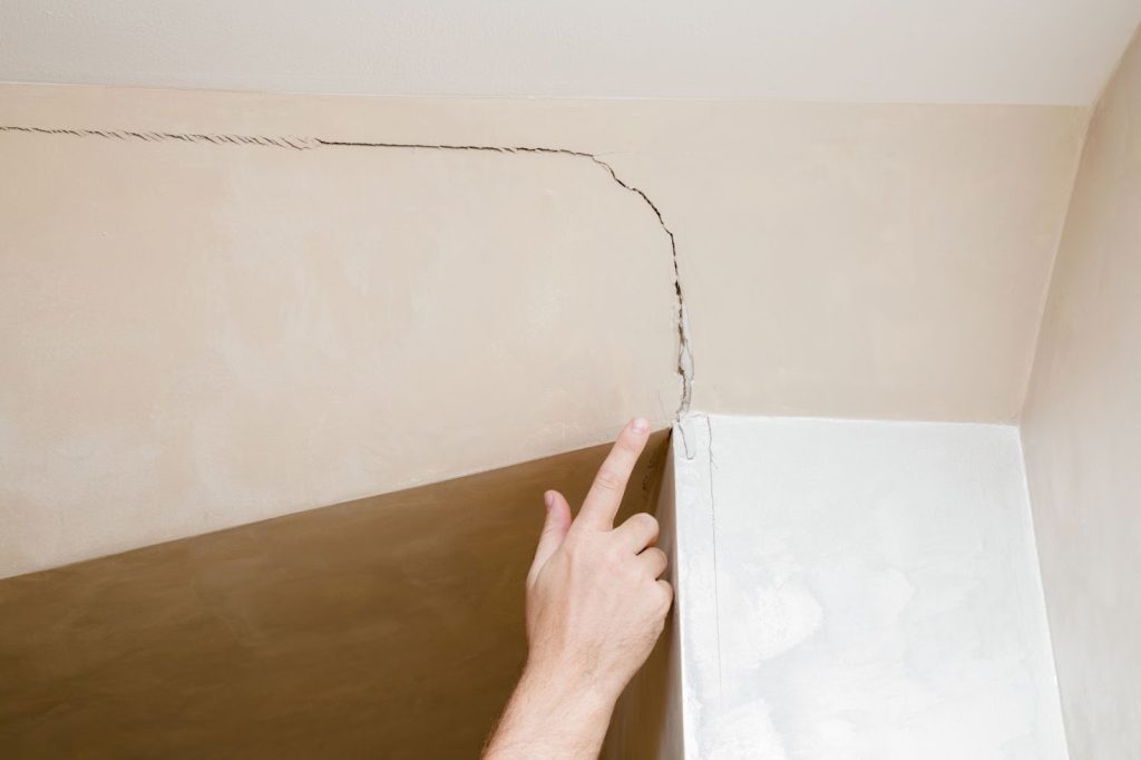 A hand pointing to a crack in the wall of a room, indicating foundation damage and the need for repair.