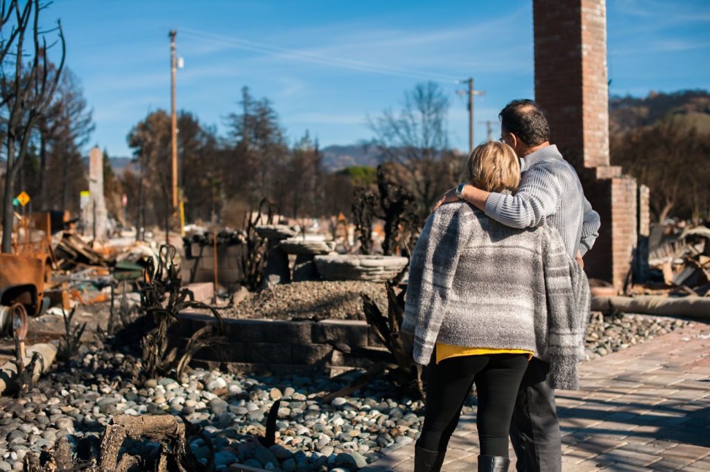 two-people-embracing-amidst-the-ruins-of-a-fire-destroyed-area-with-charred-debris-and-a-brick-chimney
